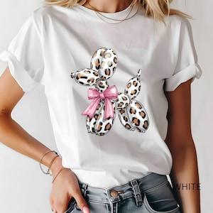 Puede incluir: Una camiseta blanca de manga corta con un gráfico central de un perro hecho de globos con estampado de leopardo y un lazo rosa brillante atado al cuello. La camiseta tiene cuello redondo y se muestra con las mangas ligeramente remangadas.