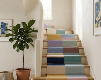 May include: A multi-coloured stair runner with a patchwork design. The runner features rectangular blocks of various colours, including red, yellow, blue, and green. The stairs are made of wood and have a white banister. A potted plant and artwork are also visible.