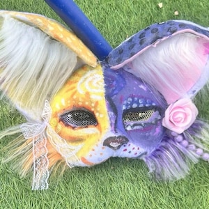 Könnte beinhalten: Eine zweifarbige Tiermaske mit einer gelben Seite aus weißem Kunstfell und Spitzenbesatz sowie einer violetten Seite mit dunklen Flecken, rosa Kunstfell, einer rosa Rose und einem kleinen Stern. Die Maske hat Netz-Augenabdeckungen und einen blauen Stab.