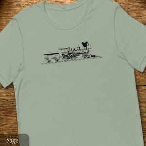 May include: Sage green t-shirt featuring a black illustration of a vintage steam train on tracks. The train is detailed with wheels, a smokestack, and a tender car. The shirt has a crew neck and short sleeves.