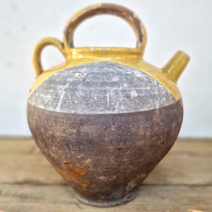 Może przedstawiać: Okrągły ceramiczny dzban z żółtą, szkliwioną górną częścią i ciemnobrązowym, teksturowanym korpusem. Naczynie posiada duży, łukowaty uchwyt u góry, mniejszy uchwyt boczny oraz krótki, ukośny dzióbek wystający po prawej stronie naczynia.