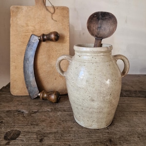 May include: A rustic kitchen scene featuring a wooden cutting board, a curved blade with a wooden handle, and a speckled ceramic jar with two handles. A wooden spoon rests inside the jar. The items are arranged on a wooden surface.