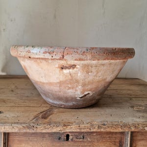 Può includere: Una ciotola in terracotta rotonda e usurata con un bordo largo e spesso. La superficie esterna presenta una patina maculata color marrone e beige terroso con crepe, scheggiature e segni visibili del tempo, incluse piccole irregolarità.