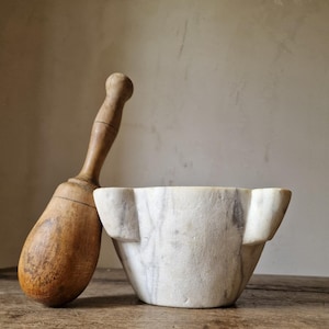 May include: A vintage mortar and pestle set. The mortar is made of white marble with gray veining, and the pestle is made of wood. The mortar has a unique shape with protruding sides. The wooden pestle is long and has a rounded handle.