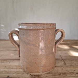Pode incluir: Um pote cilíndrico de cerâmica com um acabamento esmaltado castanho-claro liso e duas pegas curvas nos lados opostos. O recipiente apresenta um rebordo ligeiramente alargado e uma base plana, com um tom terroso consistente.