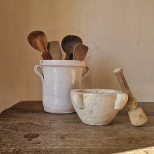 Può includere: Una scena di cucina rustica con un portautensili in ceramica bianca pieno di cucchiai di legno. Un mortaio e un pestello in pietra sono posizionati su una superficie di legno invecchiato. L'insieme è illuminato da luce naturale.