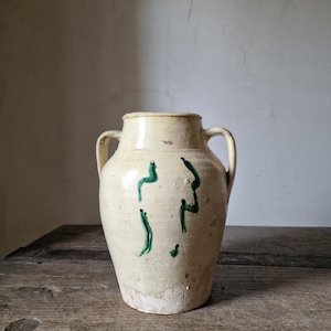 May include: A cream-colored ceramic vase with two handles and green abstract markings. The vase has a wide base and a slightly flared rim. It sits on a wooden surface.