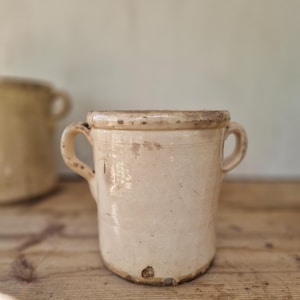 May include: A cylindrical cream-coloured ceramic pot featuring two side handles and a thick, rounded rim. The surface displays a glossy finish with visible signs of wear, including small dark spots, surface scratches, and minor chips along the base.