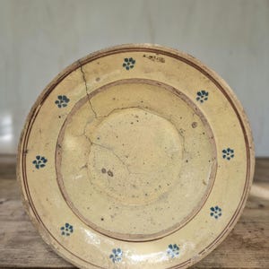 Può includere: Un piatto in ceramica circolare con finitura smaltata giallo pallido, caratterizzato da una serie di piccoli motivi floreali blu disposti lungo il bordo. Il piatto presenta una crepa evidente che si estende dal bordo verso il centro e linee marroni.