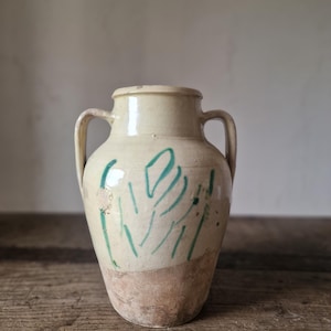 Pode incluir: Um vaso de cerâmica com duas alças laterais e um gargalo estreito. A parte superior tem um acabamento em esmalte creme com marcas lineares verdes pintadas à mão, enquanto a base inferior mantém um acabamento em terracota sem esmalte.