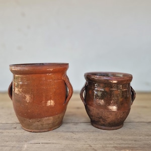 May include: Two rustic earthenware pots with glossy brown glaze and side handles. The larger pot features a lighter, unglazed base, while the smaller pot displays a darker, mottled brown finish. Both vessels have wide, circular openings and rims.