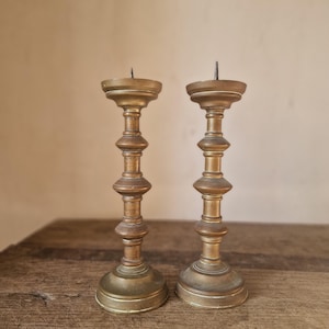 Puede incluir: Dos candeleros antiguos de latón. Presentan un diseño escalonado, con una base ancha, un tallo delgado con secciones redondeadas y una copa poco profunda para una vela. El latón tiene una pátina cálida y envejecida.