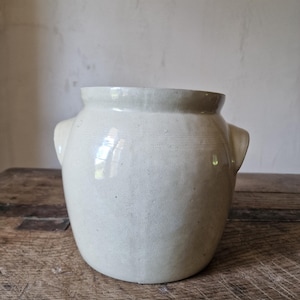 May include: A cream-colored ceramic crock with a smooth, glossy glaze and a wide, open top. The vessel features two small, integrated side handles and a rounded, bulbous body shape, showcasing a simple, functional design with subtle surface texture.