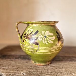 Puede incluir: Una jarra de cerámica verde de forma redondeada con un patrón floral pintado a mano en blanco y marrón oscuro. El recipiente tiene un asa lateral, un borde ancho y detalles de líneas horizontales en la base con un acabado desgastado.