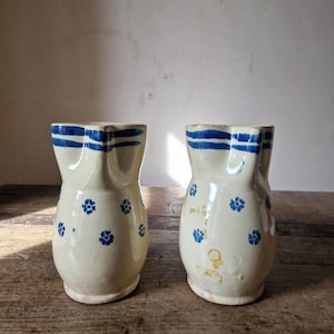 Op de afbeelding: Twee crèmekleurige keramische kannen met een handgeschilderd blauw bloemmotief en horizontale blauwe strepen rond de bovenrand. Elke kan heeft een afgerond lichaam, een uitlopende voet en een duidelijke schenktuit.