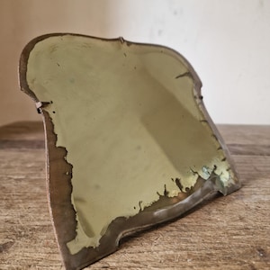 Könnte beinhalten: Ein Tischspiegel mit geschwungener, unregelmäßiger Form und einem Metallrahmen. Die reflektierende Oberfläche zeigt Alterserscheinungen mit sichtbarer Oxidation und dunklen, verwitterten Rändern entlang des Umfangs des Glases.