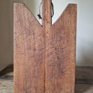 Pode incluir: Uma tábua de corte retangular de madeira castanha com uma borda superior entalhada e uma extensão de pega vertical central. A superfície apresenta um desgaste extenso com numerosas marcas de riscos visíveis e um fio no cabo.