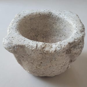 Puede incluir: Un pequeño mortero blanquecino con una superficie rugosa y texturizada. El mortero tiene un cuenco ancho y poco profundo y un borde grueso e irregular. El exterior muestra un patrón moteado y la forma general es ligeramente irregular.