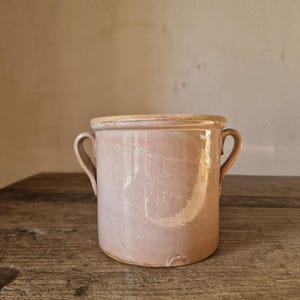 May include: A cylindrical, light pink ceramic pot with two small handles. The pot has a slightly textured surface and a light-colored rim. The pot is sitting on a wooden surface.