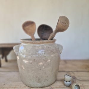 Może przedstawiać: Beżowy ceramiczny garnek z dwoma uchwytami po bokach i poziomymi żebrowaniami przy krawędzi. Wewnątrz garnka znajdują się trzy drewniane łyżki. Przed naczyniem ceramicznym leżą trzy małe, gładkie, zaokrąglone obiekty przypominające kamienie.