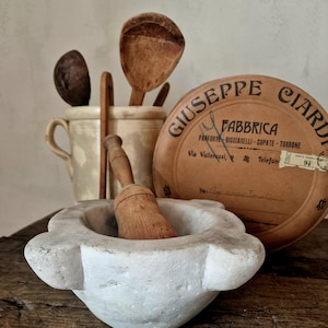 Könnte beinhalten: Ein weißer Marmor-Mörser und Stößel-Set mit einem Holzstößel. Dahinter befindet sich ein Keramikbehälter mit Holzlöffeln. Eine runde, braune Schachtel mit italienischem Text deutet auf ein Vintage-Küchen-Thema hin.