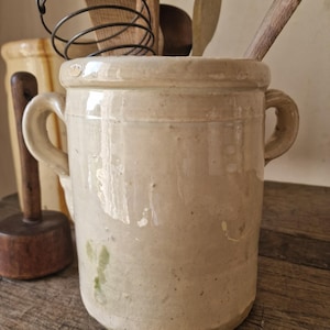 Puede incluir: Un recipiente para utensilios de cocina de cerámica de color crema, de estilo antiguo, con dos asas. El recipiente contiene cucharas de madera y un batidor de metal. Tiene un aspecto ligeramente desgastado. El fondo incluye otros utensilios de cocina.