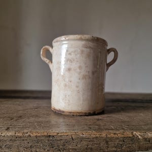 May include: A cylindrical ceramic crock featuring a cream-colored glaze with visible brown mottled staining and weathering. The vessel has two small, curved handles on opposite sides and a thick, rounded rim, showing signs of age and wear.