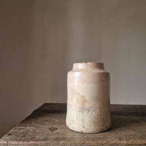 Op de afbeelding: Een cilindervormig steengoed vat met een beige, gevlekte glazuurlaag en een licht uitlopende rand. Het oppervlak heeft een zichtbare textuur, subtiele kleurvariaties en een verweerd uiterlijk met een duidelijke horizontale band.