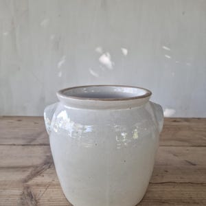 Puede incluir: Una vasija de cerámica de color crema con un esmalte suave y brillante y una boca ancha. El recipiente cuenta con dos pequeñas asas laterales integradas y un borde superior marrón sin esmaltar, mostrando un diseño sencillo y funcional.