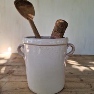 May include: A cylindrical off-white ceramic crock with two side handles, containing a dark brown wooden spoon and a weathered, textured brown wooden pestle. The ceramic surface features a glossy finish with subtle, small indentations throughout.