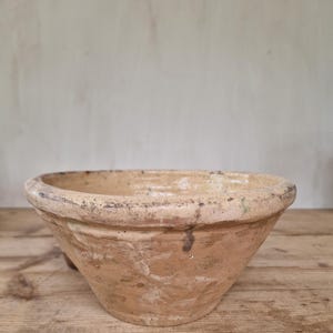 Può includere: Una ciotola in terracotta con bordo largo e base conica. L'esterno presenta una superficie ruvida color marrone chiaro con macchie scure. L'interno ha una finitura liscia e smaltata in tonalità beige crema con leggere variazioni di colore.