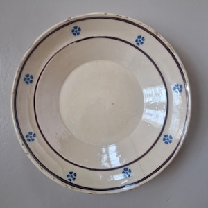 Op de afbeelding: Een rond, gebroken wit keramieken bord met een donkerbruine cirkelvormige band rond het midden. De rand is versierd met zes kleine, blauwe, vijfbladige bloemmotieven die gelijkmatig over de omtrek van de brede, platte rand zijn verdeeld.