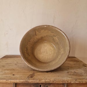 May include: A round, deep ceramic bowl with a mottled, light beige and tan glazed interior. The exterior of the bowl features a smooth, solid beige finish with a slightly darker rim. The bowl has a wide, open top and a curved, basin-like shape.
