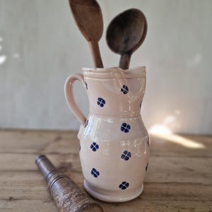 Może przedstawiać: Jasnoróżowy ceramiczny dzbanek z powtarzającym się wzorem małych ciemnoniebieskich czteropłatkowych kwiatów. Wewnątrz dzbanka znajdują się dwie ciemnobrązowe drewniane łyżki, a na pierwszym planie leży poziomo cylindryczny drewniany tłuczek.