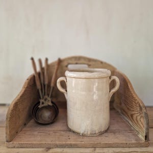 Pode incluir: Um pote de cerâmica cor de creme com duas alças laterais posicionado ao lado de um conjunto de copos medidores de metal com cabos de madeira, tudo dentro de um suporte de madeira curvo com um recorte central para alça e veios visíveis.