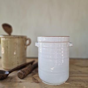May include: A cylindrical white ceramic crock featuring a glossy finish, horizontal ribbed detailing along the body, and two small side handles. The vessel has a wide opening at the top and a flat base, presenting a clean and functional design.