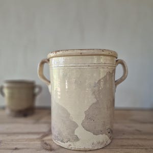 May include: A cylindrical ceramic crock with two side handles, featuring a cream-colored glazed finish with visible areas of wear and exposed brown clay patches. The vessel has a thick rim and a smooth, slightly tapered body design.