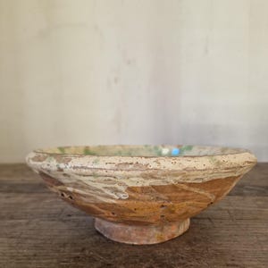 May include: A rustic, antique ceramic bowl with a wide, shallow design. The bowl features a weathered appearance with a mix of brown, beige, and green hues. The bowl has a small, round base and a textured surface.