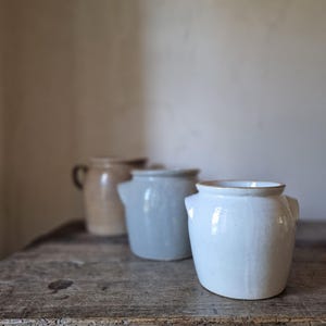 Puede incluir: Tres vasijas de cerámica con asas laterales dispuestas en fila. La vasija delantera es blanca, la del medio es azul claro y la trasera es de color tostado. Cada recipiente presenta un cuerpo cilíndrico con un borde ligeramente ensanchado.