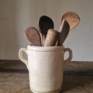 以下が含まれることがあります： 2つのサイドハンドルが付いたクリーム色の陶器製クロック。中には、複数のスプーンや質感のある木製のすりこぎ棒など、さまざまな木製のキッチン用品が円筒形の容器の中に立てて収納されています。