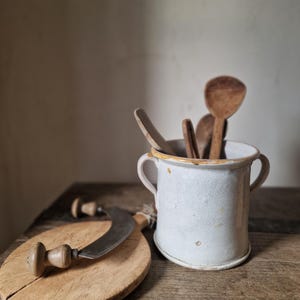 May include: A white ceramic crock with two side handles containing several wooden spoons. Next to the crock sits a curved metal mezzaluna blade with wooden handles resting on a round, light-coloured wooden cutting board with a visible crack.