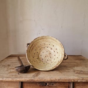 May include: A cream-colored ceramic colander with two side handles and a series of drainage holes, accompanied by a dark brown wooden ladle. The colander features a textured, aged finish with visible variations in the glaze and surface color.