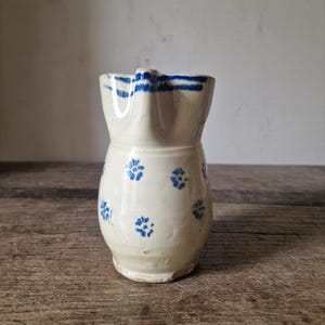 Op de afbeelding: Een crèmekleurige keramische kan met een herhalend patroon van kleine blauwe bloemmotieven en horizontale blauwe strepen rond de rand. De kan heeft een afgerond lichaam, een uitlopende bovenkant en een duidelijke schenktuit aan de voorzijde.