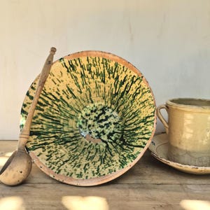 Puede incluir: Un cuenco de cerámica grande y redondo de color amarillo con un patrón de esmalte verde, acompañado de un cucharón de madera de mango largo y una taza de cerámica amarilla lisa con dos asas laterales sobre un plato a juego, todo dispuesto junto.
