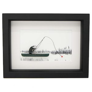 May include: Framed artwork featuring a whimsical illustration of a figure fishing from a small boat. The artwork is in black and white with a touch of red, set against a white background. The black frame adds a clean, modern look.