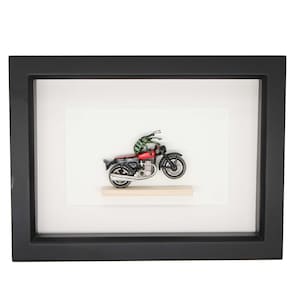 May include: A framed artwork featuring a toy motorcycle with a green and black insect figurine riding on it. The motorcycle is red, black, and silver. The artwork is set against a white background.