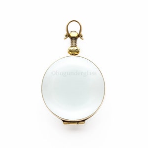 May include: A gold locket with a hinged lid and a round, clear glass window. The locket has a decorative, ornate top with a loop for attaching a chain.
