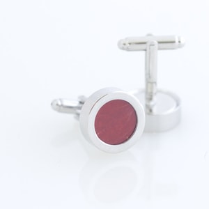 May include: A pair of silver cufflinks with a circular design. Each cufflink features a polished silver frame surrounding a deep red, textured center. The cufflinks are displayed on a white surface, highlighting their metallic sheen.