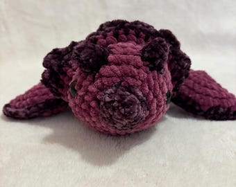May include: A handmade, plush turtle in shades of burgundy and purple. The crocheted turtle has a textured, velvety appearance, with small black eyes and flipper-like appendages. The turtle is lying on a white surface.