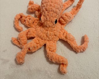 May include: A handmade, peach-colored crochet octopus toy. The plush octopus has eight textured tentacles and small black eyes. The soft, cuddly toy is made of a thick, looped yarn.